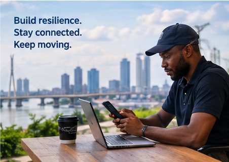 A man working on a laptop and phone at an outdoor table with a city skyline behind him — build resilience, stay connected, keep moving
